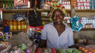 En kvinder sidder smilende blandt varer i sin købmandsbutik