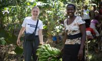 En frivillig ung kvinde fra Operation Dagsværk bærer smilende en bananklase gennem en plantage sammen med en ugandisk frivillig