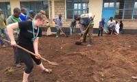 Mange mennesker, blandt andet en spejder med tørklæde og Spejderhjælpen t-shirt, skovler jord i Rwanda