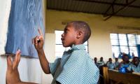 En dreng i skoleuniform løser regnestykker ved tavlen i et klasselokale med bliktag i Rwanda